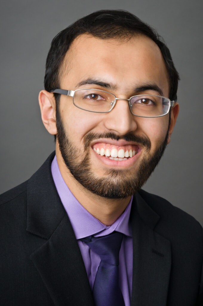 Headshot of a man in his 20s with heritage from Pakistan with light-medium skin tone, short black hair, and short black beard. He is wearing a black suit with purple button up shirt and navy tie and is wearing silver thin rimmed oval glasses. He is smiling at the camera.