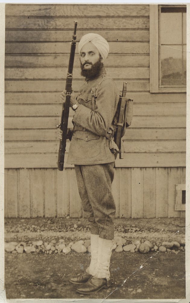 Sepia tone photograph of a man in a military uniform with a white turban and large beard, he is holding a rifle and his backpack has a bayonet.  