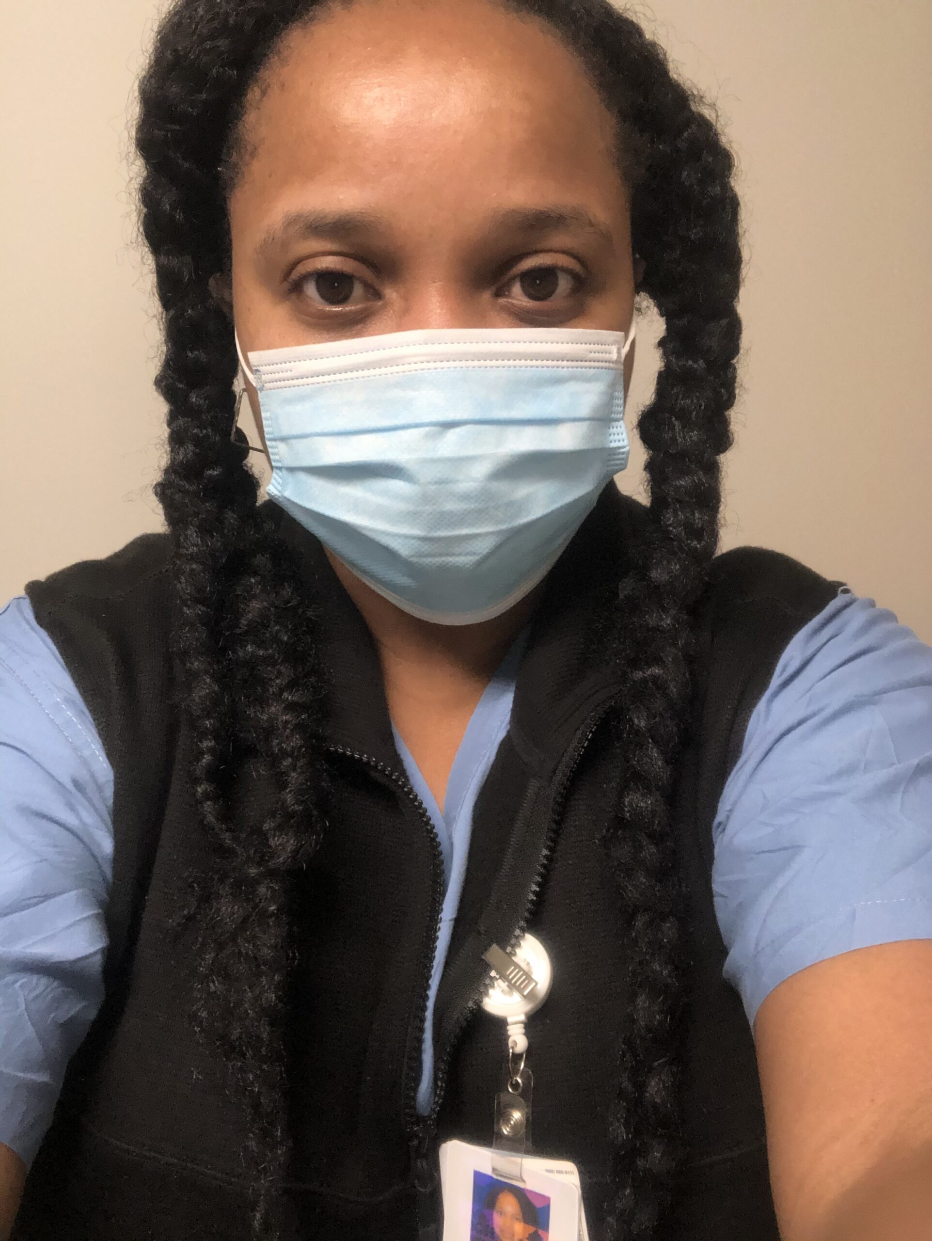 Selfie of a woman in her 20s who has heritage from Nigeria, with medium skin tone and long black braided hair. She is wearing a surgical mask, blue scrubs and black fleece vest, with an ID badge.