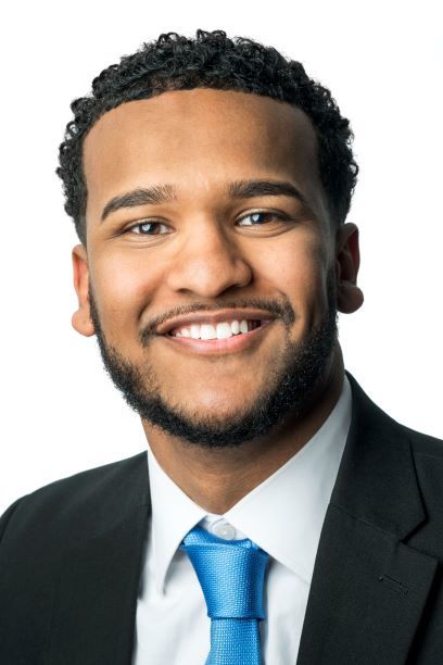 Headshot of a man in his 20s who has heritage from Kenya with medium skin tone, short curly black hair and a short beard. He is wearing a black suit, white button up and blue tie. He is smiling at the camera. 