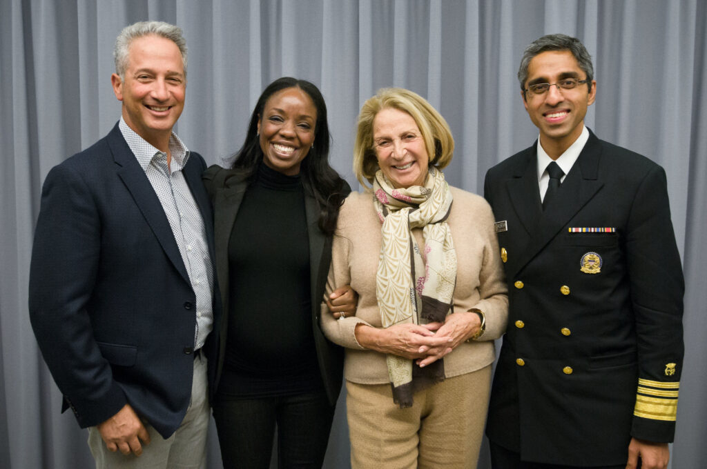 Photograph of four people posing and smiling for the camera. On the left a man in his 50s with light skin tone and grey short hair. Next a woman in her 40s with medium-dark skin tone and straight black hair. Next a woman in her 80s with light skin tone and chin length blonde hair. On the right a man in his 40s with medium skin tone and dark grey hair, he is wearing a military uniform. 