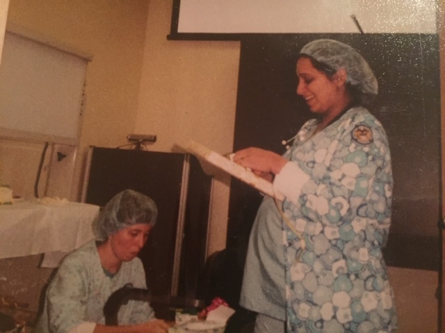 Photograph of two women both in scrubs and hairnets. One woman crouches over a baby car seat. The other woman stands with a clipboard in hand.