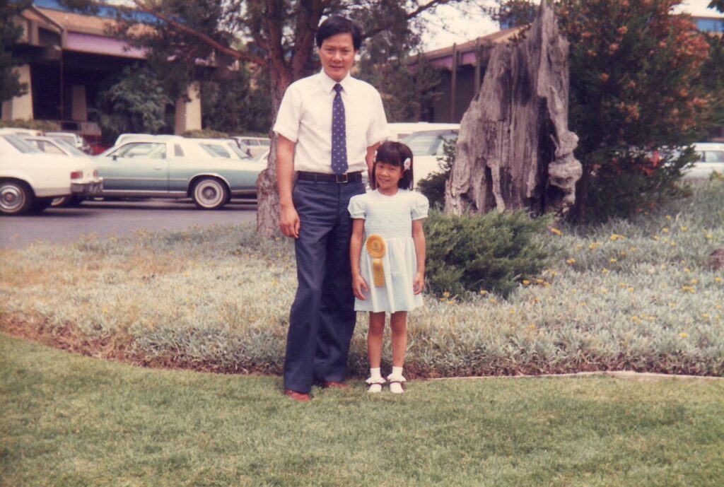 Old photograph of a man in his 30s wearing a button up short sleeve, blue slacks and a blue tie, he stands with his hand on the back of a young girl wearing a blue dress and has a yellow ribbon pined to her waist.