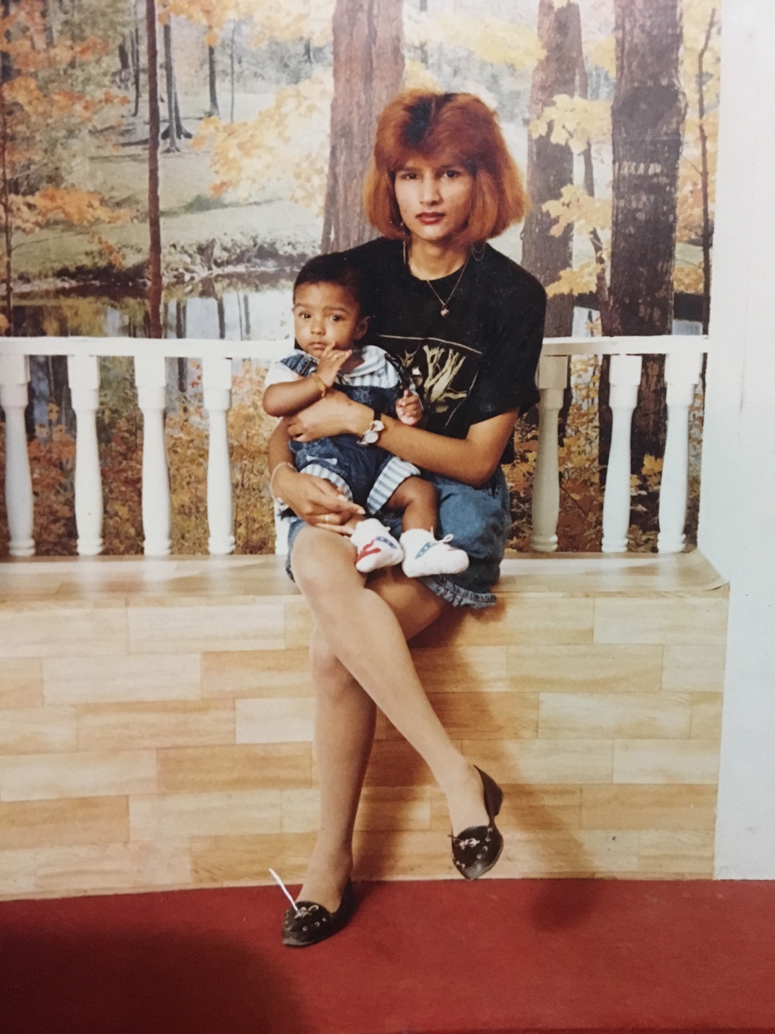 Old photograph of a woman in her 20s with light skin tone and dyed red teased hair. She is seated on a bench, with a railing and photograph of trees behind her. She is holding a baby.