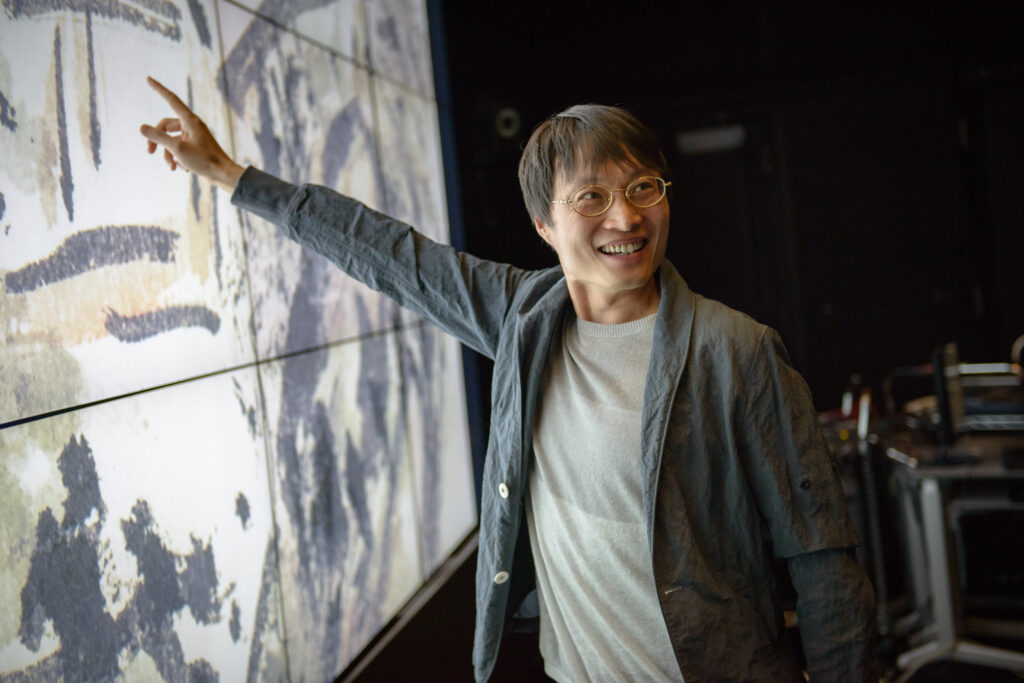 Photograph of a man in his 40s who has heritage from China with light skin tone and black hair, he is wearing a grey jacket, light grey sweater and gold oval glasses. He is looking off camera and pointing in the opposite direction at a screen. 