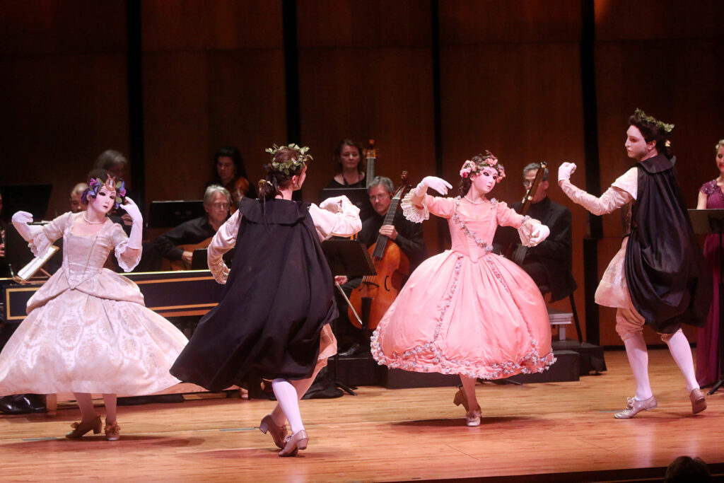 Photograph of a performance. Two people in costume dance in the foreground, behind them musicians perform.