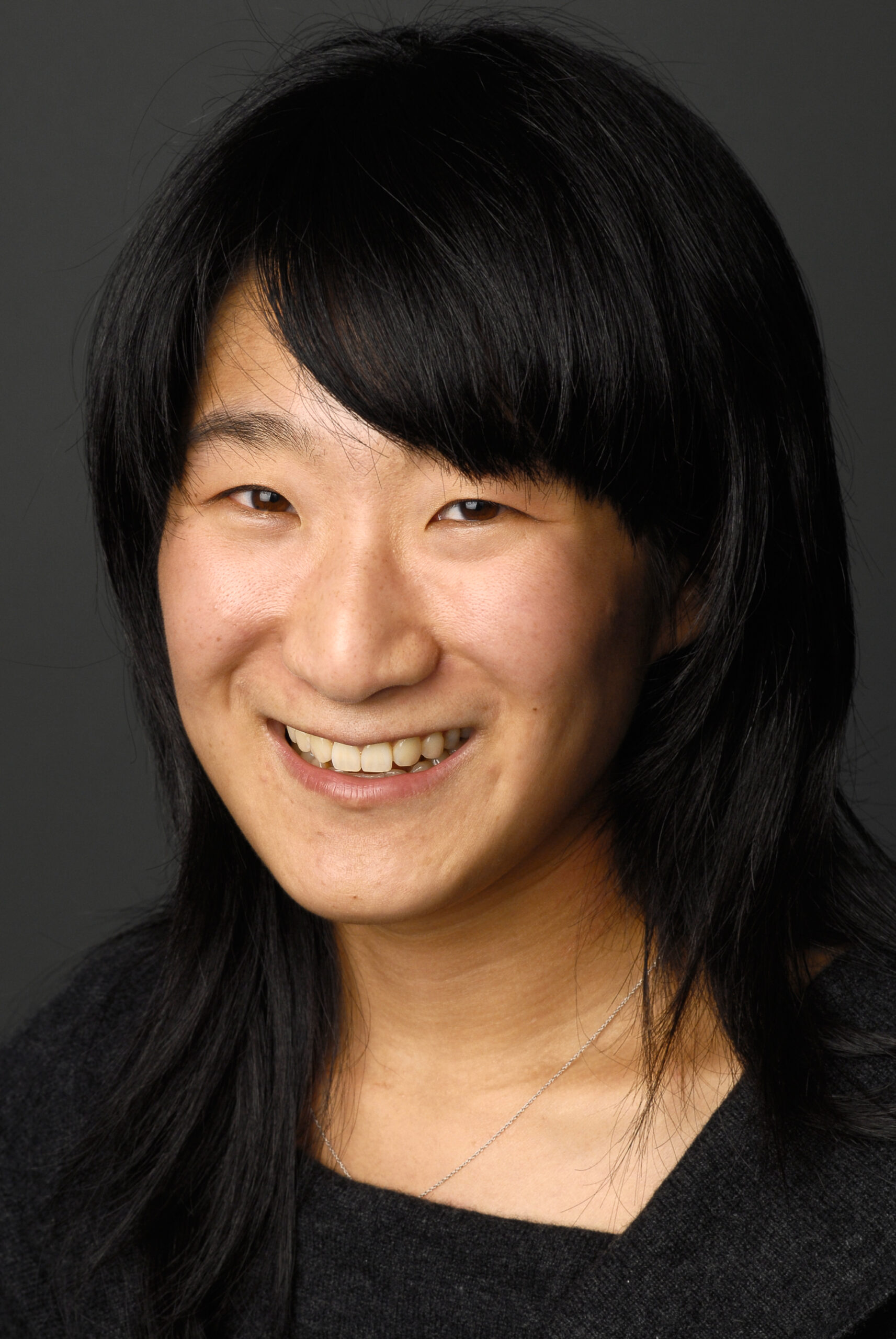 Headshot of a woman in her 30s with heritage from South Korea, she has medium-light skin tone and black shoulder length straight hair with swooped bangs. She is wearing a black top and smiling at the camera.