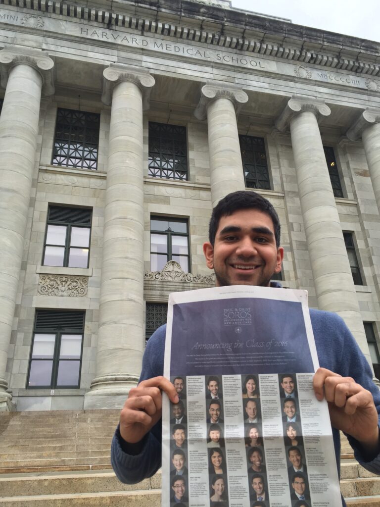 Photograph of a man in his 20s with light skin tone and short black hair. He is wearing a blue top, standing in front of a large stone building with columns and holding a New York Times. 