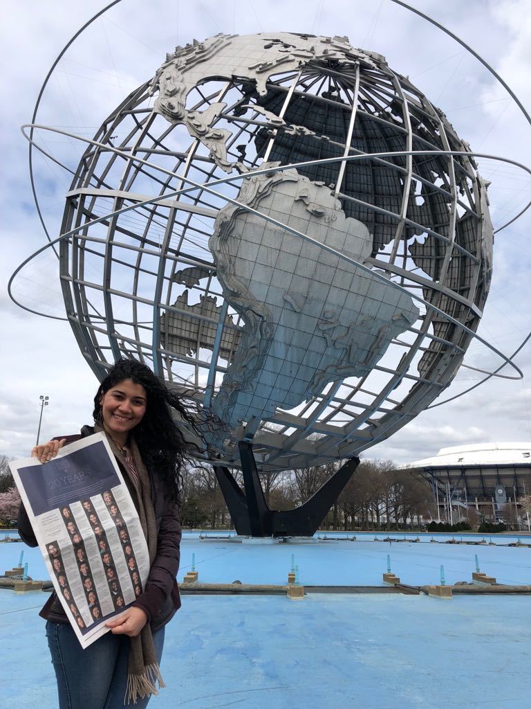 Photograph of a woman in her 20s who has heritage from Colombia with light skin tone and black hair. She is holding a New York Times and standing in front of the Unisphere in Flushing Meadows - Corona Park, Queens.