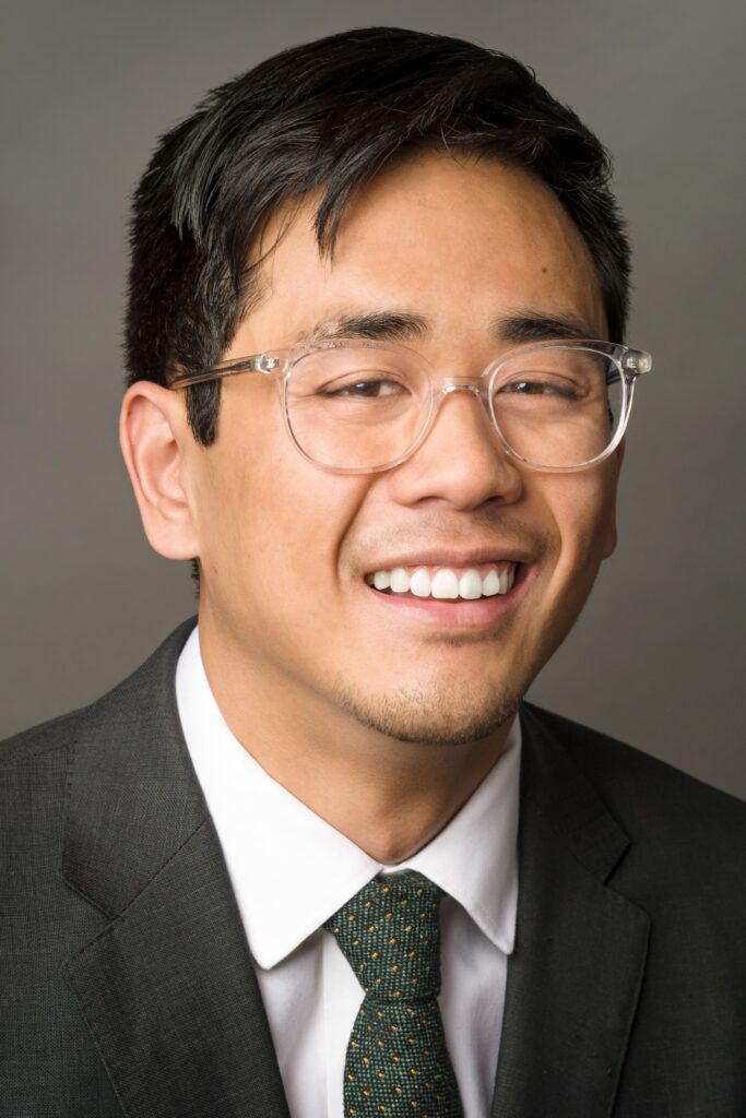 Headshot of a man in his 20s who has heritage from Cambodia with medium skin tone and black combed over hair. He is wearing a grey suit, white button up shirt and green tie, plus clear rimmed large glasses. He is smiling at the camera. 