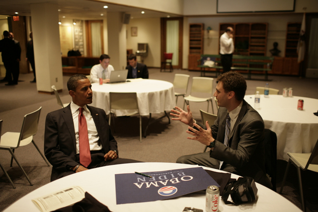 Photograph of two men sitting at a round table with a white table cloth. The man on the left has medium skin tone and buzzed black hair, he is wearing a black suit, white button up shirt and red tie. The man on the right has light skin tone, brown short hair and is wearing a grey suit, white button up shirt and multi-colored tie, he is gesturing and the two appear in deep conversation. 