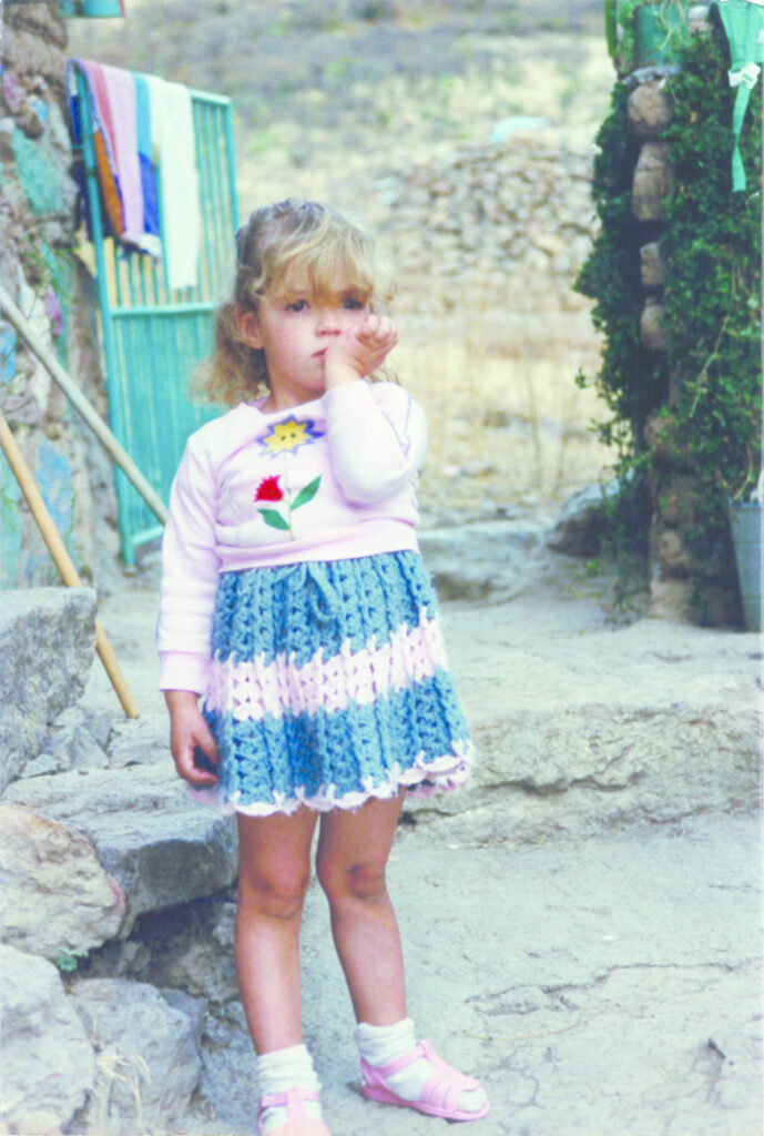 Photograph of a young girl with light skin tone and blonde hair, wearing a pink long sleeve with a flower on it, and a crocheted blue and pink skirt.