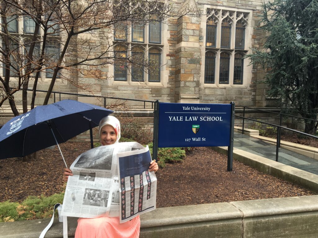 Photograph of a woman in her 20s who has light skin tone and is wearing a white hijab. She is holding an umbrella in one hand and the New York Times open in the other. She sits next to a sign that reads "Yale Law School"