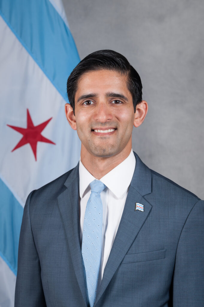 Headshot of a man in his 30s who has heritage from India with light skin tone and short black hair. He is wearing a blue suit, white button up shirt and light blue tie, there is a Chicago flag pin on his lapel. 