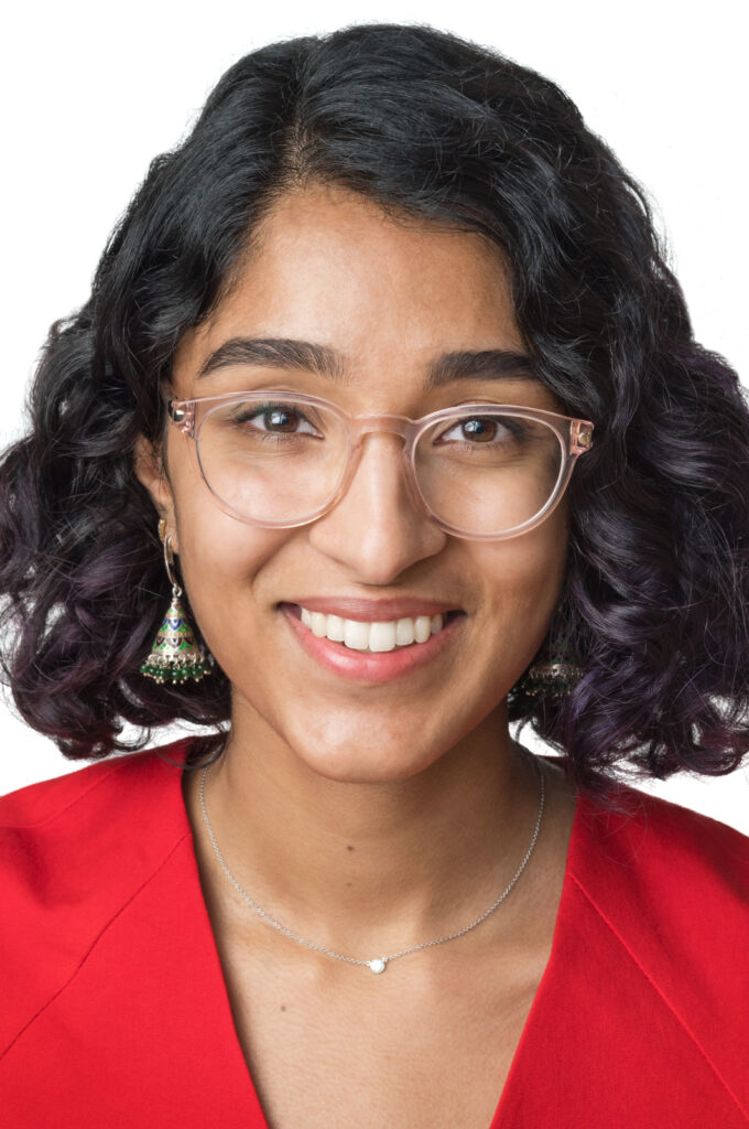 Headshot of a woman in her 20s who has heritage from India, with medium skin tone and chin length black curly hair, parted on the right side. She is wearing a red blouse, silver necklace and multi-colored pyramid shaped earrings, and light translucent large-framed glasses. She is smiling at the camera.  