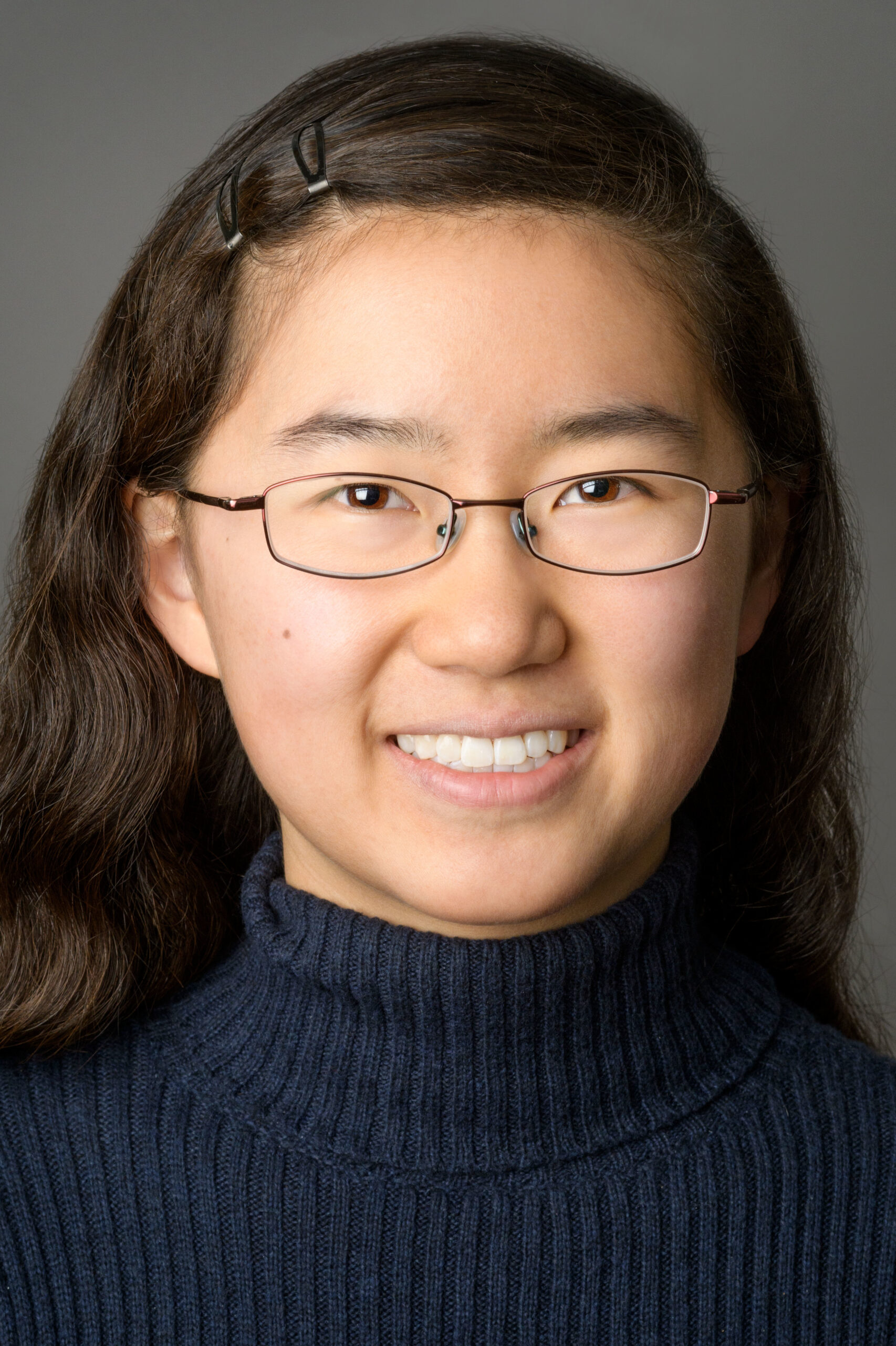 Headshot of a woman in her 20s who has heritage from China with light skin tone and wavy long dark brown hair, parted on the far left with two snap clips on the right. She is wearing a navy blue turtleneck sweater and small oval glasses. She is smiling at the camera.