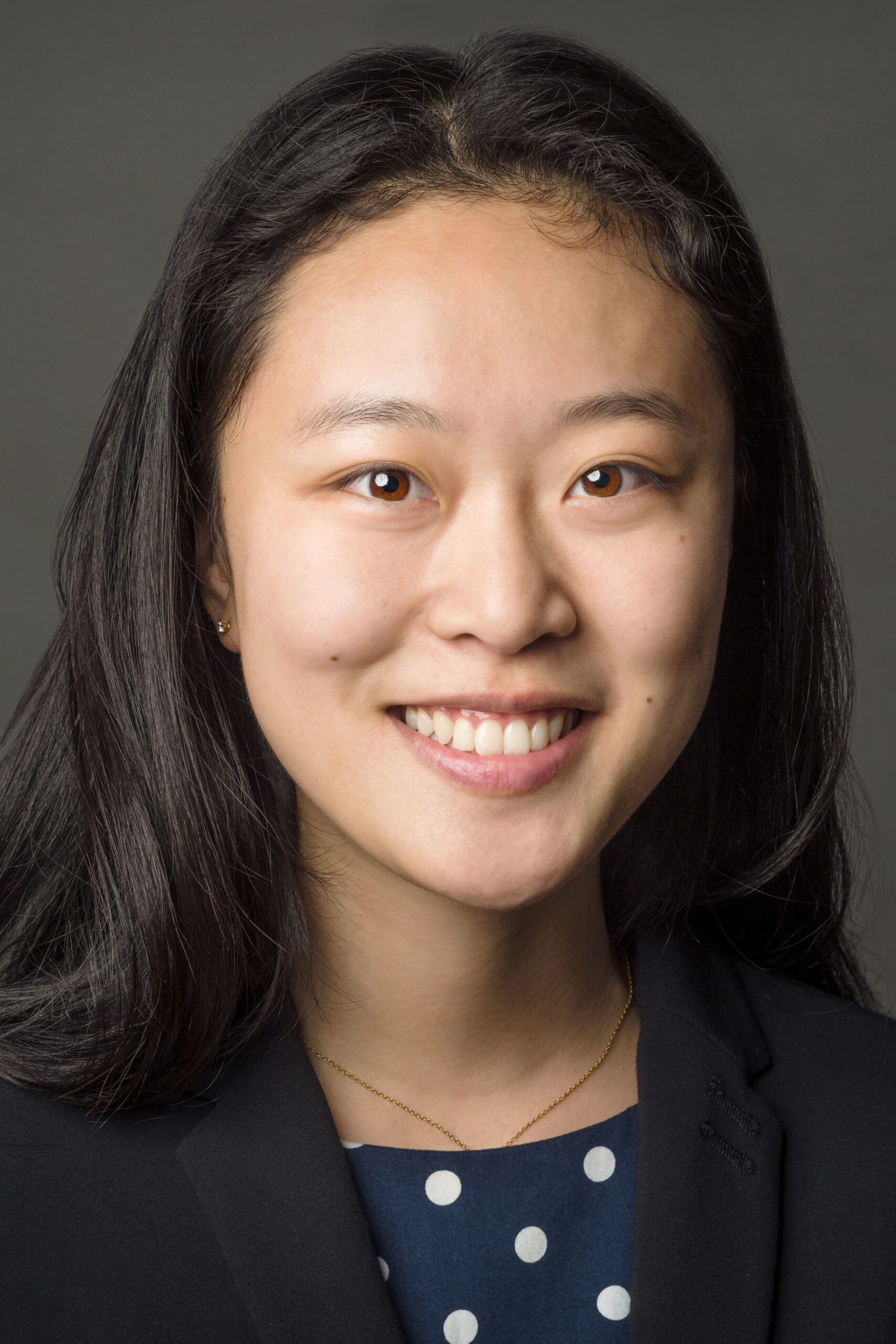 Headshot of a woman in her 20s with heritage from China who has light skin tone and long black hair. She is wearing a black blazer with a blue with white polka dot blouse, gold necklace and stud earrings. She is smiling at the camera. 