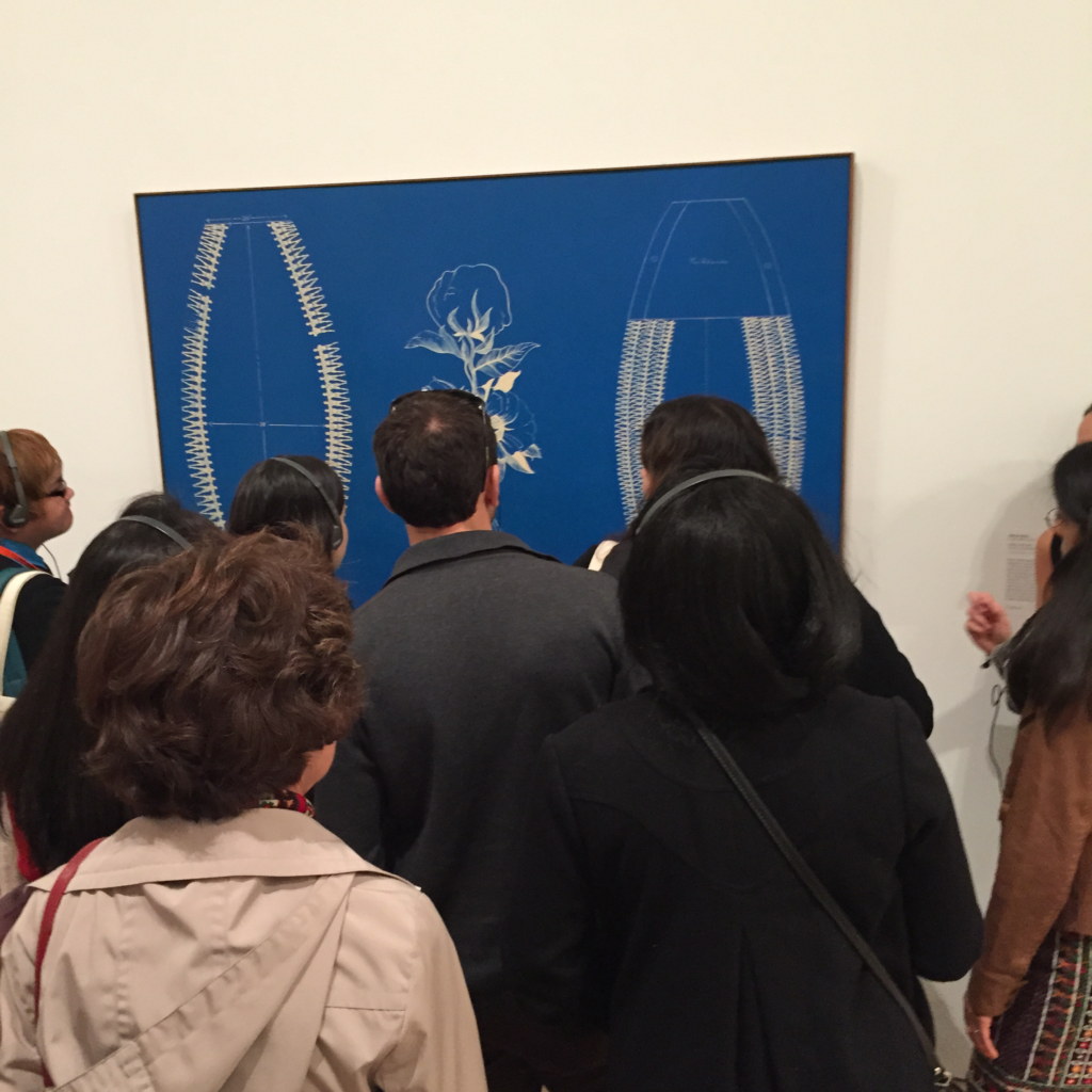 Photograph from the back of the crowd of people looking at the same piece of art. It is bright blue with white. 