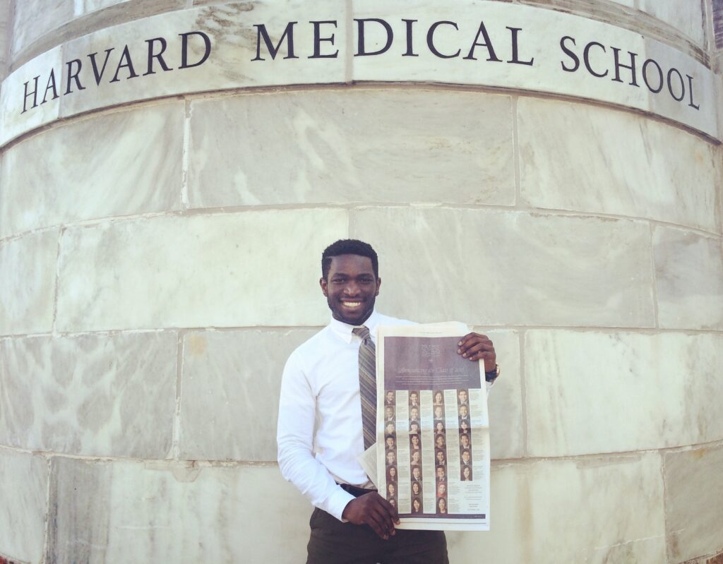 Photograph of a man in his 20s with dark skin tone, short black hair and a trim beard, he is wearing a white button up shirt and tie. He stands in front of a stone wall that says "Harvard Medical School" and holds the New York Times. 