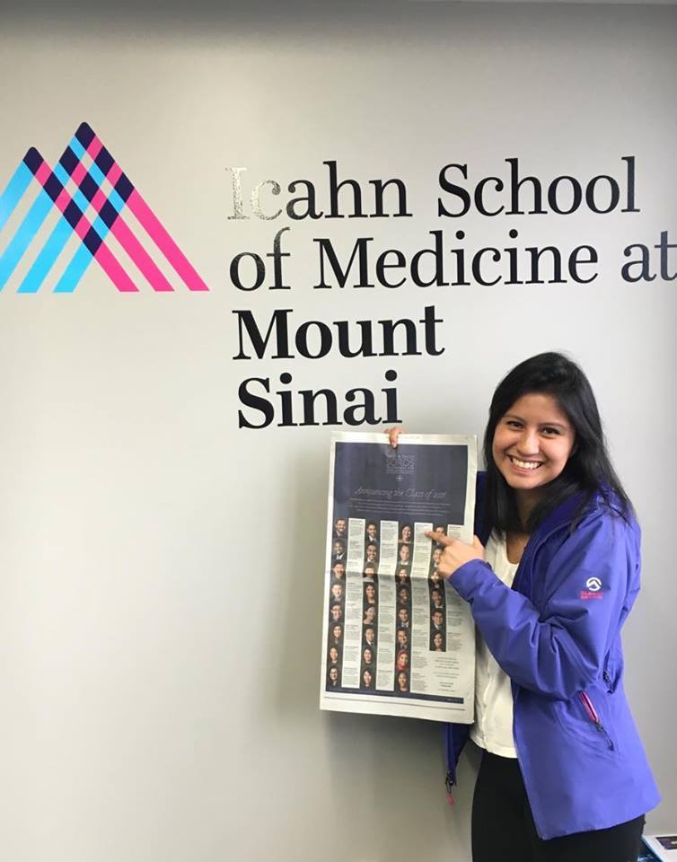 Photograph of a woman in her 20s with light skin tone and long black straight hair. She is wearing a purple jacket and holding the New York Times under a sign that says Icahn School of Medicine at Mount Sinai. 