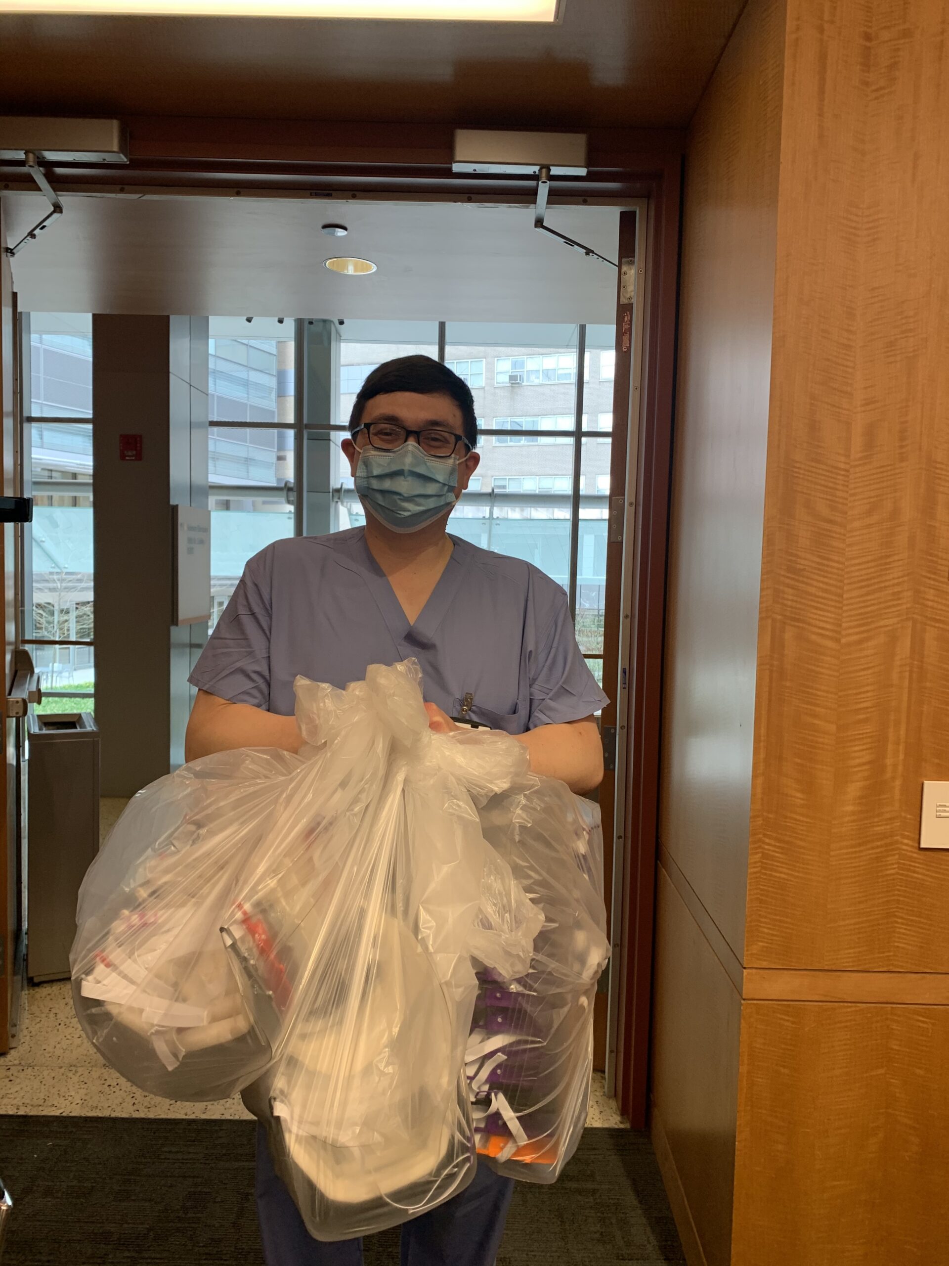 Photograph of a man in his 40s with heritage from Indonesia and Czech Republic. He is wearing blue scrubs, a surgical mask and black rimmed oval glasses. He is standing in a doorway holding three large bags of face shields. 