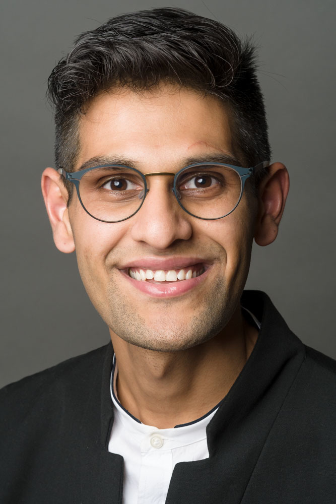 Headshot of a man in his 20s who has heritage from India, he has medium skin tone and crew cut black hair. He is wearing a black eastern collar suit and white shirt, he has thin framed round glasses. He is smiling at the camera. 