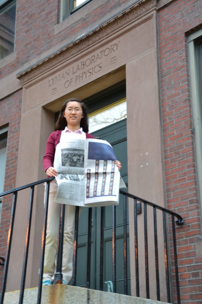 Photograph of a woman in her 20s who has heritage from China, with light skin tone and long wavy black hair. She is standing at a railing under a doorway that reads "Lyman Laboratory of Physics". She is holding a New York Times.