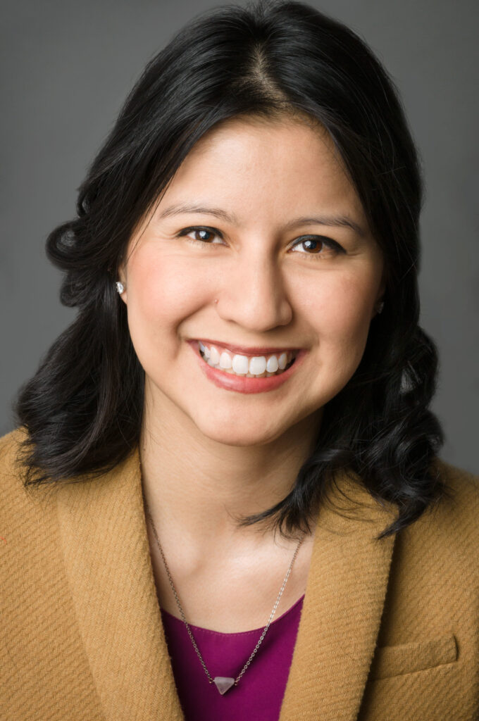 Headshot of a woman in her 20s who has heritage from Mexico, she has light skin tone and shoulder length, black curled hair. She is wearing a mustard yellow blazer, pink blouse, pink and silver necklace and stud earrings. She is smiling at the camera.