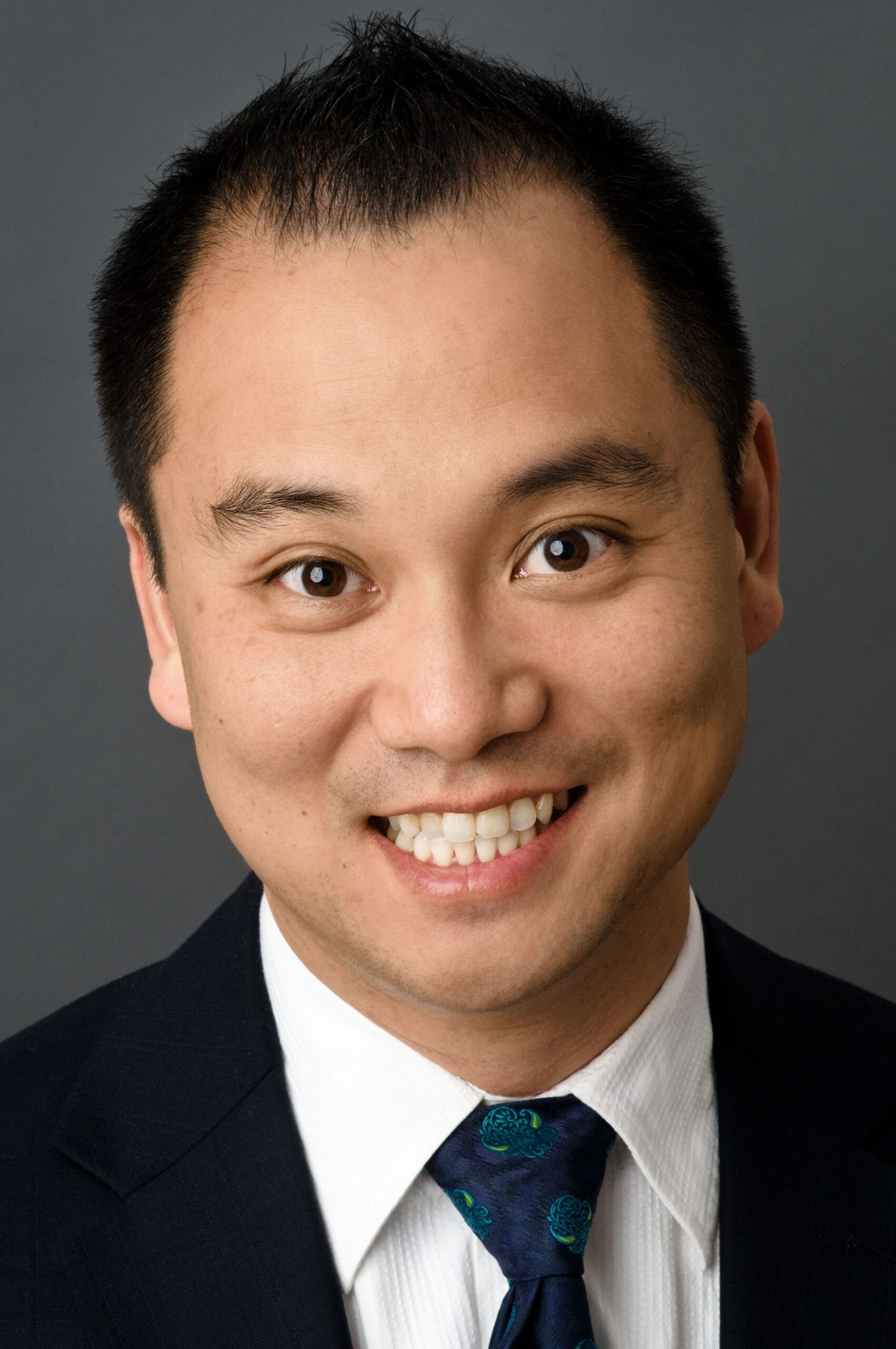 Headshot of a man in his 30s who has heritage from Hong Kong he has light skin tone and black short hair. He is wearing a black suit, white button up shirt and blue tie. He is smiling at the camera.