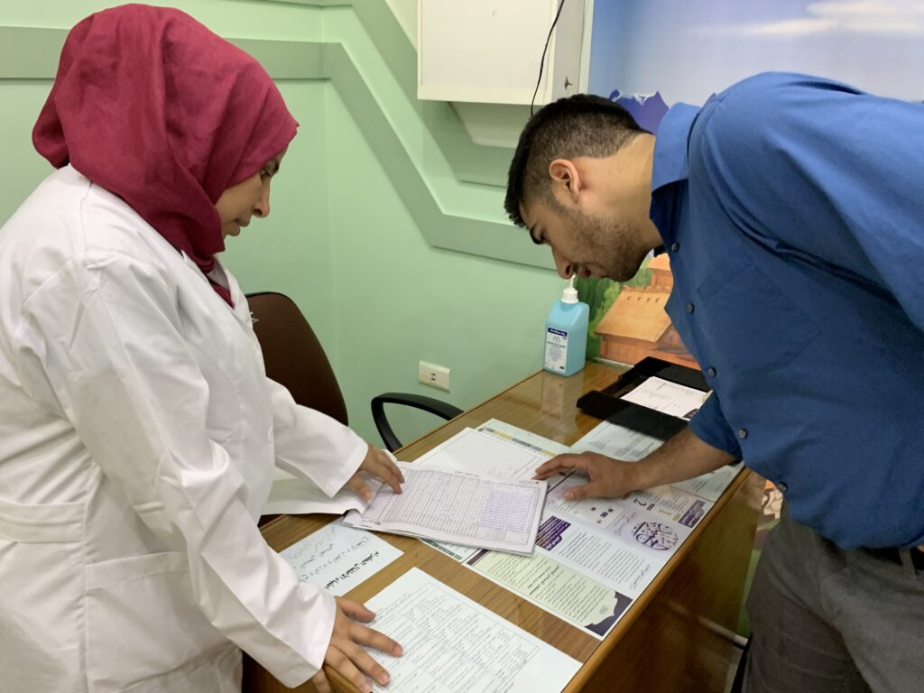 Photograph of an office, a man stands on the right in a blue button up shirt, bending over papers on the desk and reading them. A woman on the left wears a red hijab and white doctor's coat. 