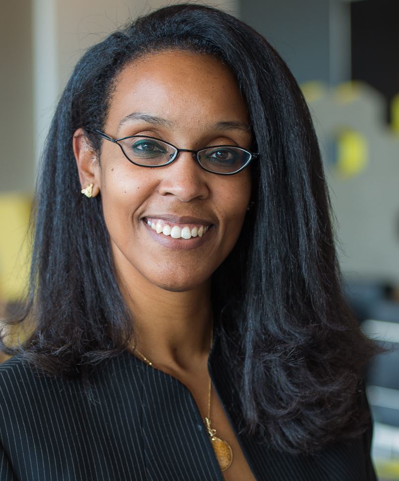 Headshot of a woman in her 30s who has heritage from Jamaica with medium-dark skin tone and shoulder length straightened black hair with a side part. She is wearing a pinstripe collarless jacket, a gold locket, gold earrings and thin framed small oval glasses. She is smiling at the camera.