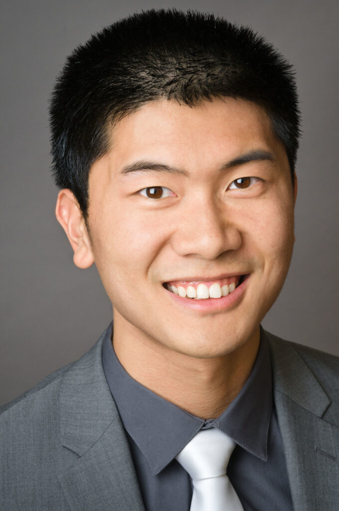 Headshot of a man in his 20s with heritage from China, he has light-medium skin tone and black short hair. He is wearing a grey suit, grey button up shirt and white tie. He is smiling at the camera. 