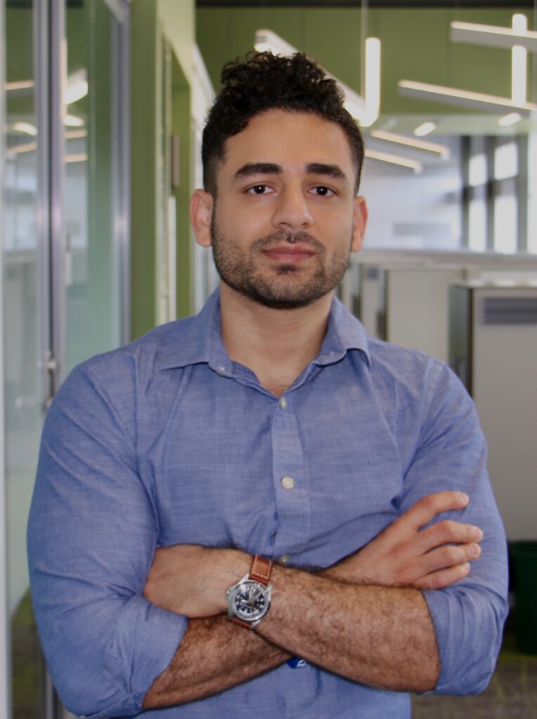 Half body photograph of a man in his 30s who has heritage from Oman with light skin tone, black crew cut hair and a trimmed beard. He is wearing a blue button up shirt and a watch with a brown leather band, his arms are crossed on his chest. 