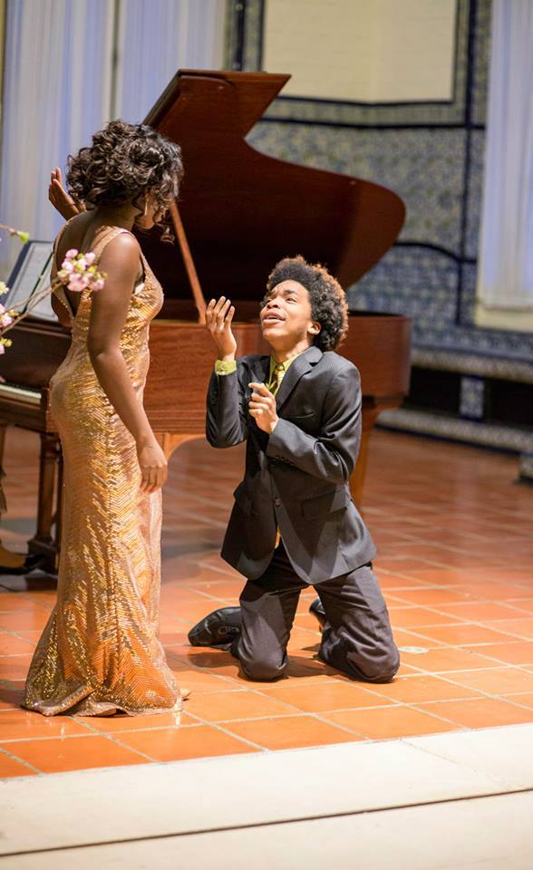 Photograph of two people standing on a stage. The woman on the left has medium-dark skin tone with black hair in an up-do, she is dressed in a gold gown. She is looking at a man with medium skin tone and black afro hair, he is on his knees gesturing toward her, he is wearing a grey suit, green button up and multi-colored tie. A piano can be seen behind them. 