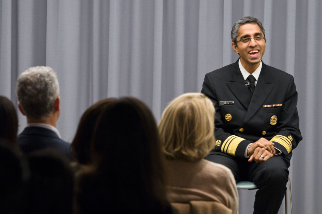 Photograph taken from a few rows back, we can see heads. A man in his 40s who has medium skin tone and short grey hair, sits on a stage with a grey curtain behind him. He is wearing a military uniform and small rimmed rectangle glasses. He is smiling at the crowd. 