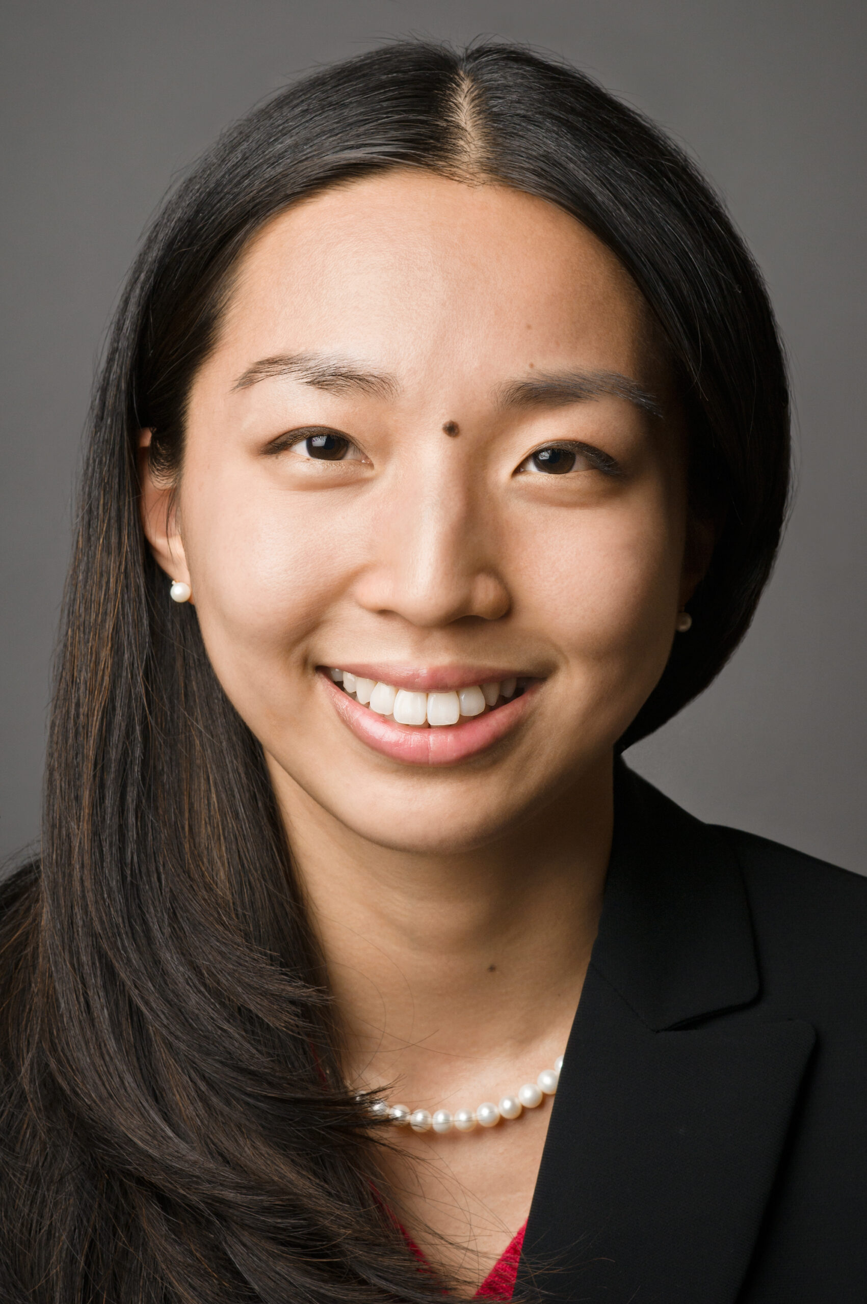 Headshot of a woman in her 20s with heritage from China who has light-medium skin tone with long, black, middle parted hair. She is wearing a black blazer, red shirt, pearl necklace and pearl stud earrings. She is smiling at the camera. 