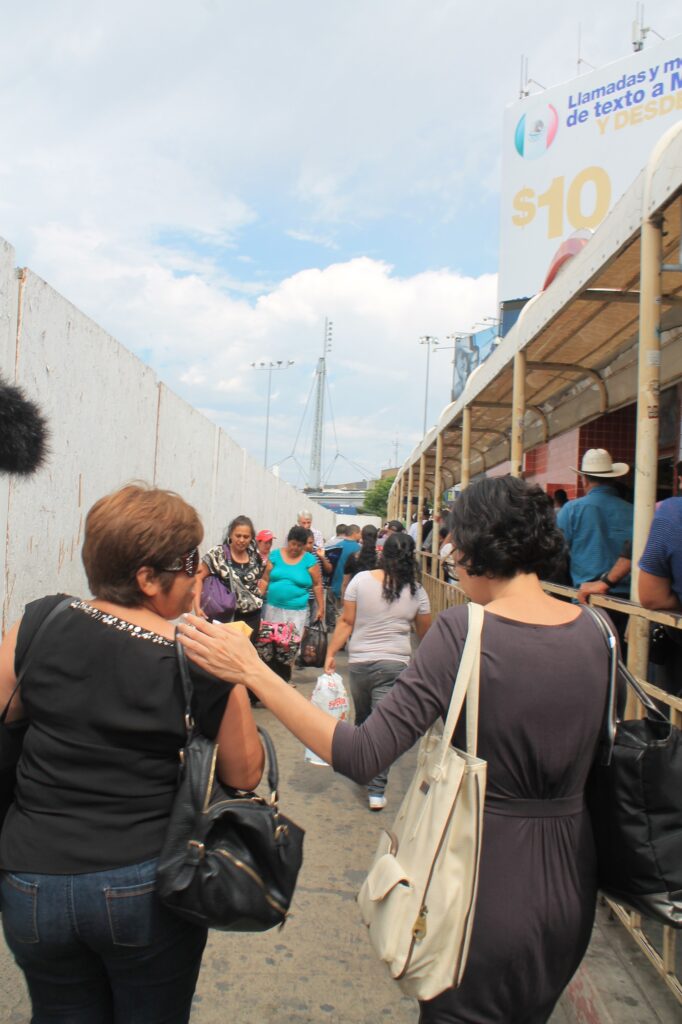 The woman on the right who has heritage from Iran has light skin tone and short black curly hair. She is wearing a grey dress and carrying a large white bag and a large black bag, one on each shoulder. She rests her left hand on a woman's shoulder next to her.