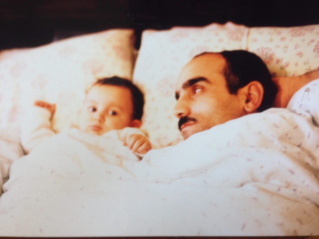 Old photograph of a man and baby tucked into white with small flower bedsheets. The man has light skin tone and black hair and a black mustache, he is looking at the baby. 