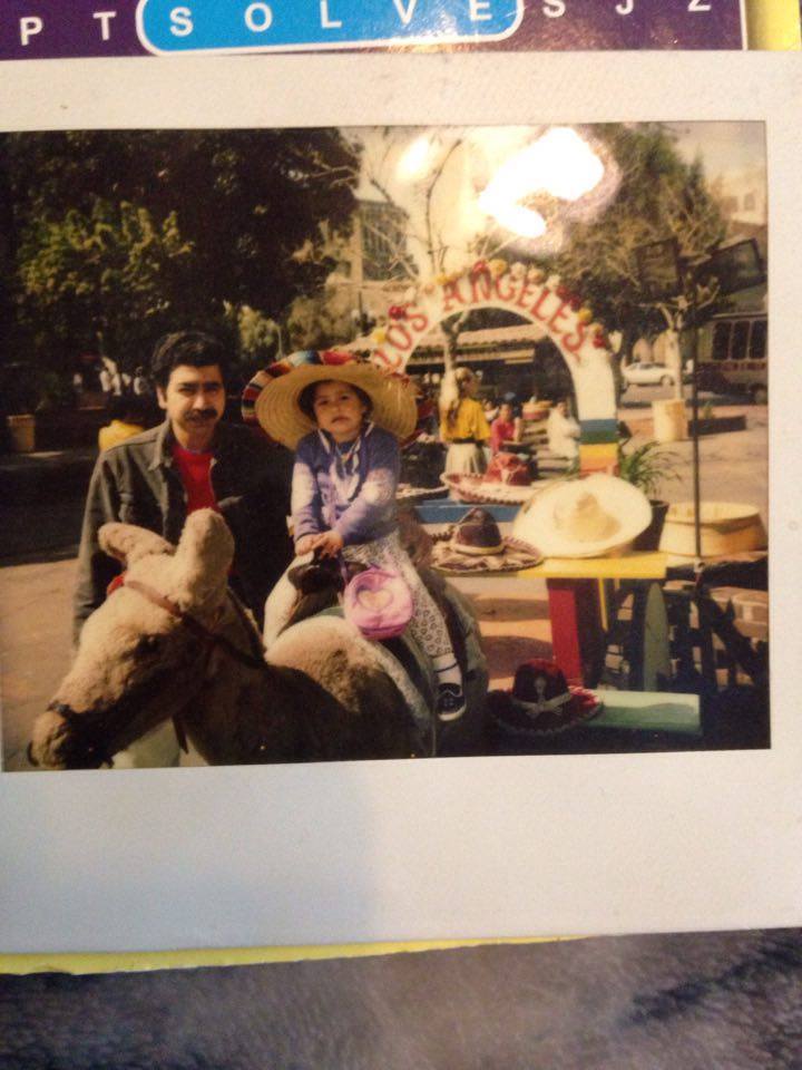 Old photograph of a man in his 20s with light skin tone, black hair and black mustache, he is wearing a red shirt and black jean jacket. He stands next to a young girl wearing a sombrero, purple long sleeve and light leggings, she has a pink bag over her shoulders.