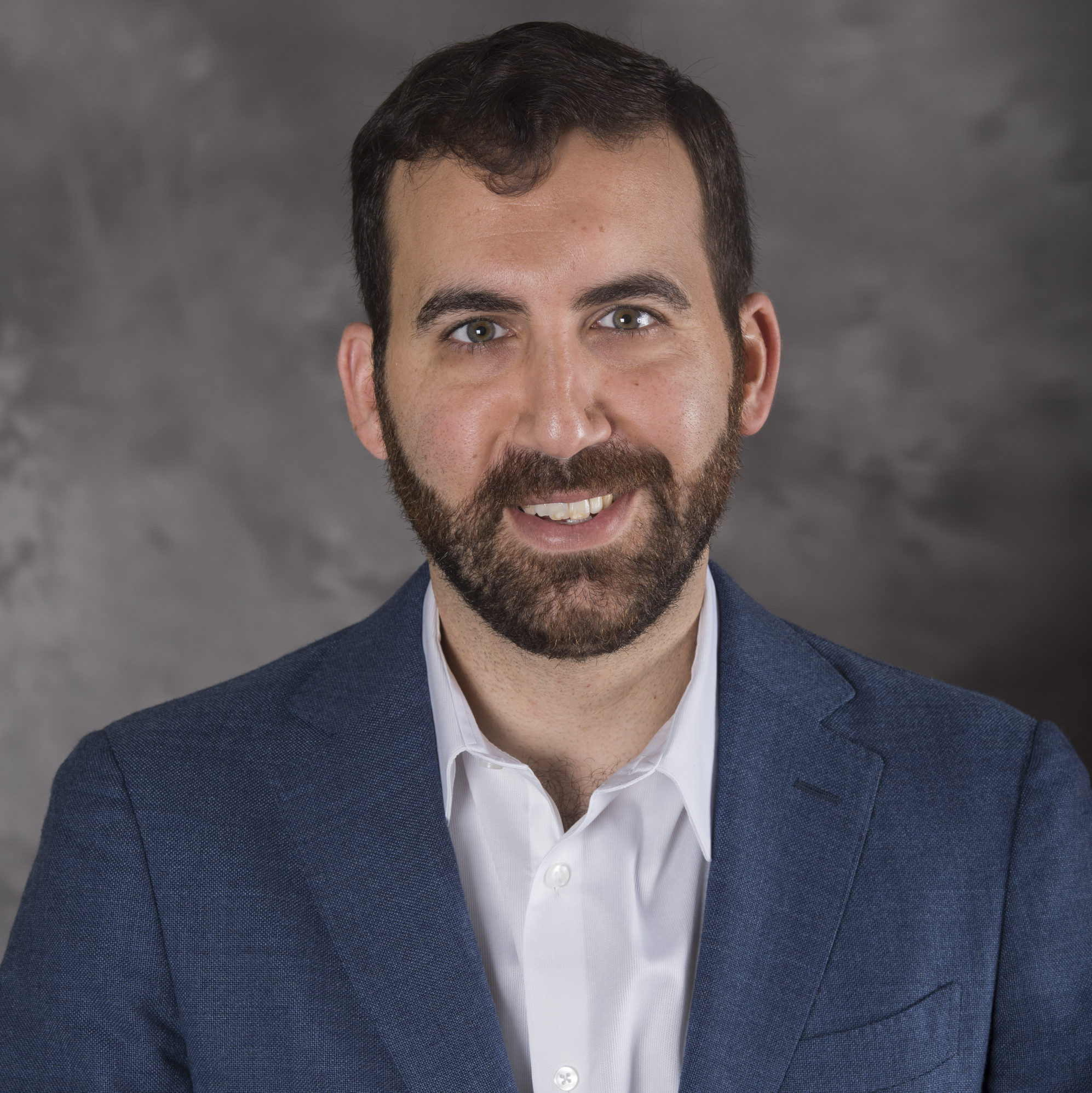 Headshot of a man in his 20s who has heritage from Lebanon and Afghanistan who has light skin tone and short dark brown hair, with a short brown beard. He is wearing a blue suit and white button up shirt. He is smiling at the camera.