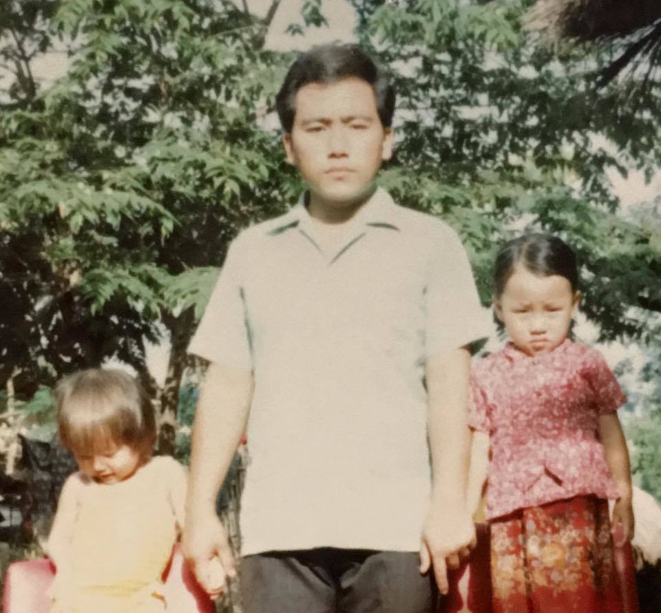 Old photograph of a man in his 30s with heritage from Thailand, with short black hair, wearing a light-colored button up tee shirt. He is holding hands with a child on each side. They are standing in front of a tree.