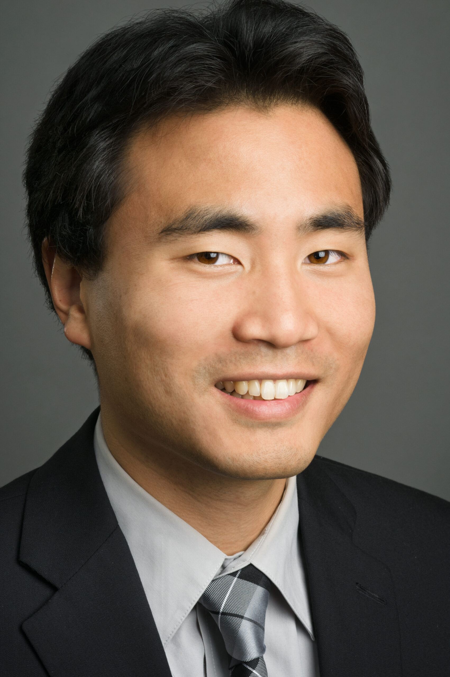 Headshot of a man in his 20s who has heritage from Taiwan and China with medium skin tone and black hair. He is wearing a black suit, light grey button up and grey tie. He is smiling at the camera.