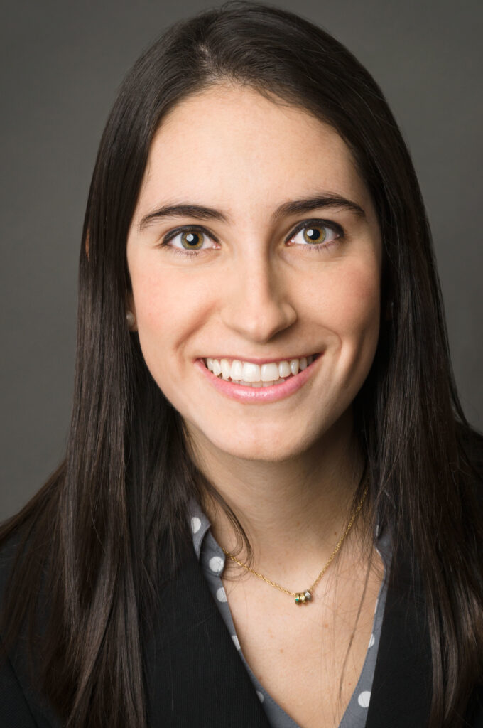 Headshot of a woman in her 20s who has heritage from Colombia, with light skin tone and long black straight hair parted on the side. She is wearing a black blazer, with grey with white polka dot button up and a gold necklace. She is smiling at the camera.