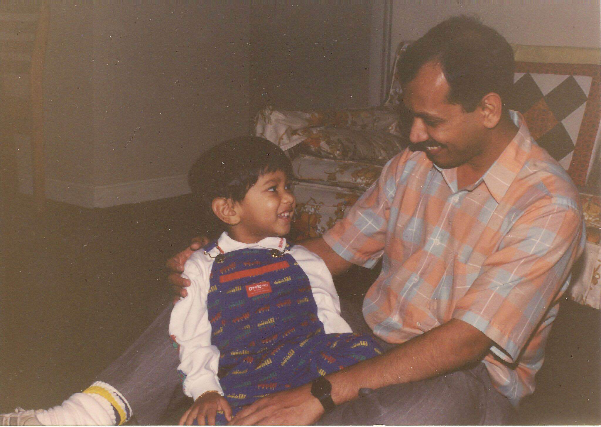 Old photograph of a man in his 30s who has medium skin tone and black hair and mustache. He is sitting on the ground engaging with a young boy. 