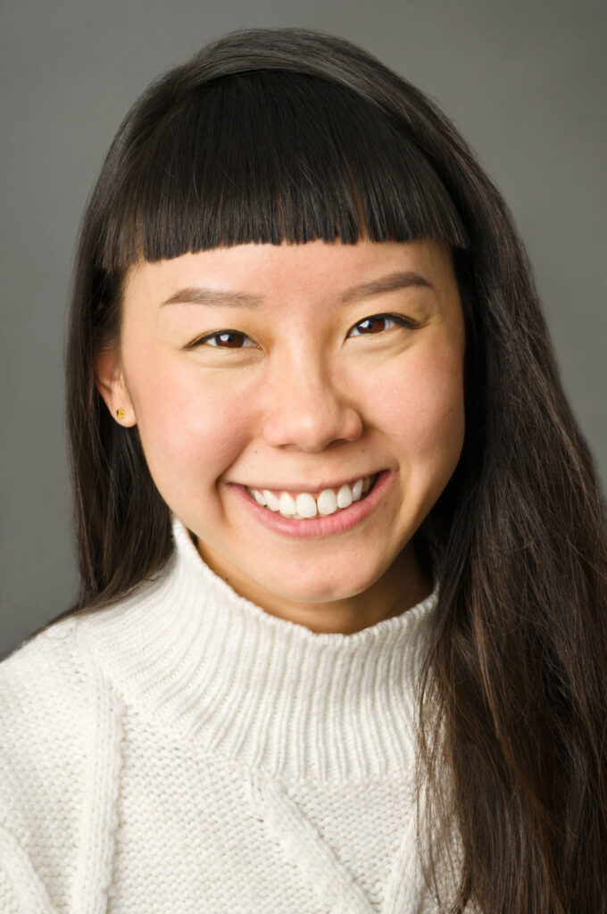 Headshot of a woman in her 20s who has heritage from Taiwan with light skin tone, long black straight hair and straight across bangs. She is wearing a white turtleneck sweater and smiling at the camera.