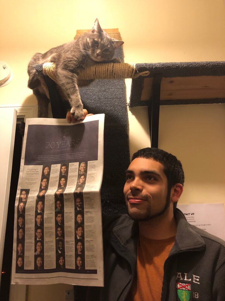 Photograph of a man in his 20s who has heritage from Mexico and Colombia with light-medium skin tone and buzzed black hair. He is holding up a New York Times so it looks as though the gray cat above him is holding the newspaper. 