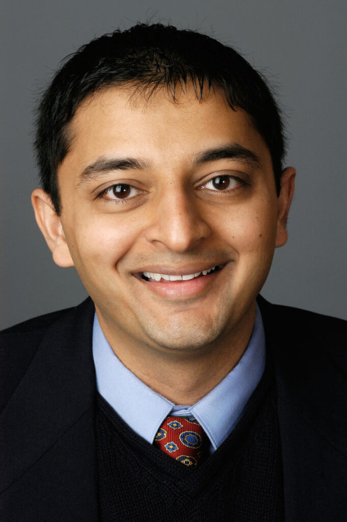 Headshot of a man in his 20s who has heritage from India with medium skin tone and black short cut hair; he is wearing a black blazer, black v-neck sweater, light blue button up shirt and a red tie, he is smiling at the camera