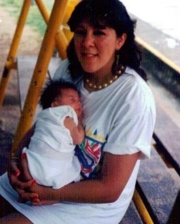 Old photograph of a woman in her 20s with light skin tone and dark long hair. She is wearing a large white t-shirt, sitting on a bench and holding a new baby. 