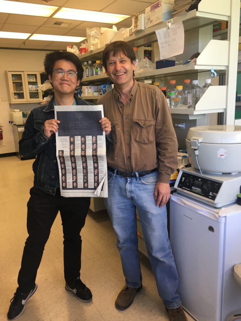 Photograph of two men standing in a lab. The man on the right is in his 40s wearing a brown button up and jeans. The man on the left is in his 20s who has heritage from South Korea with light skin tone and black hair. He is holding a New York Times.