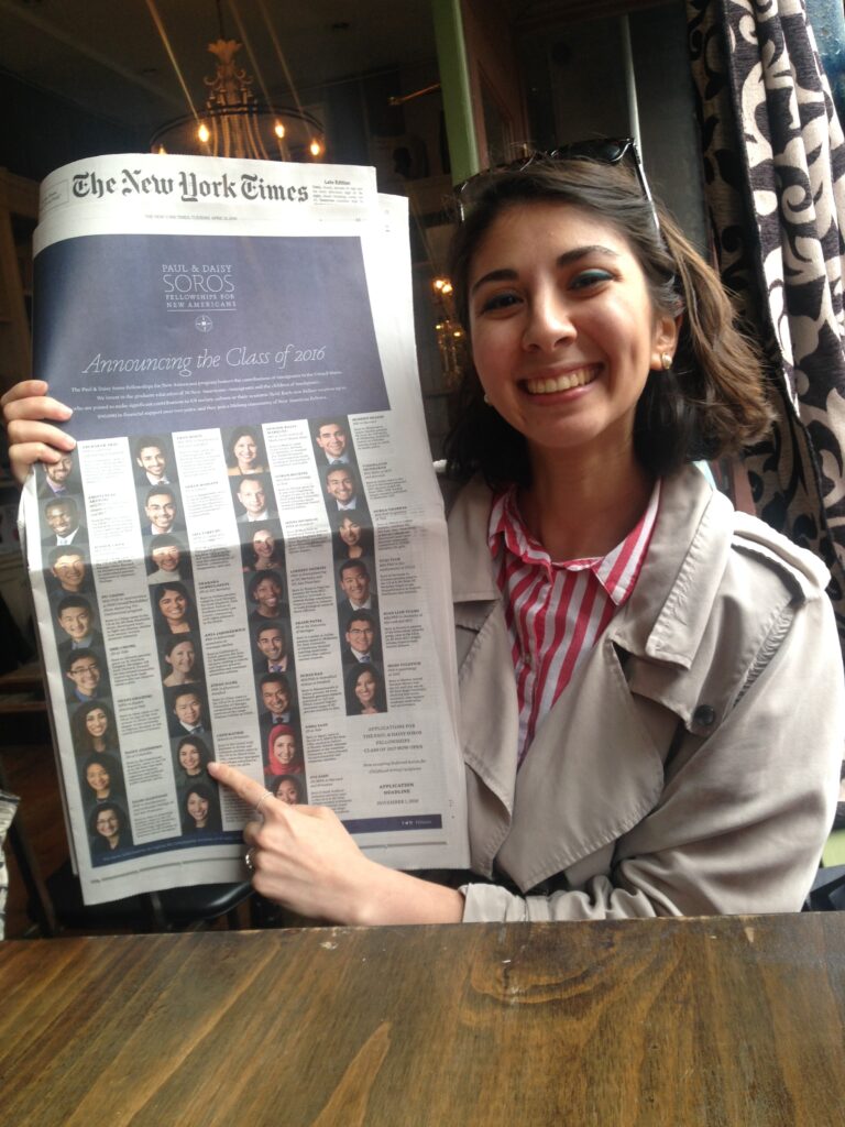 Photograph of a woman in her 20s who has light skin tone and chin length dark brown hair. She is wearing a tan jacket and red and white striped shirt underneath. She is holding up the New York Times.